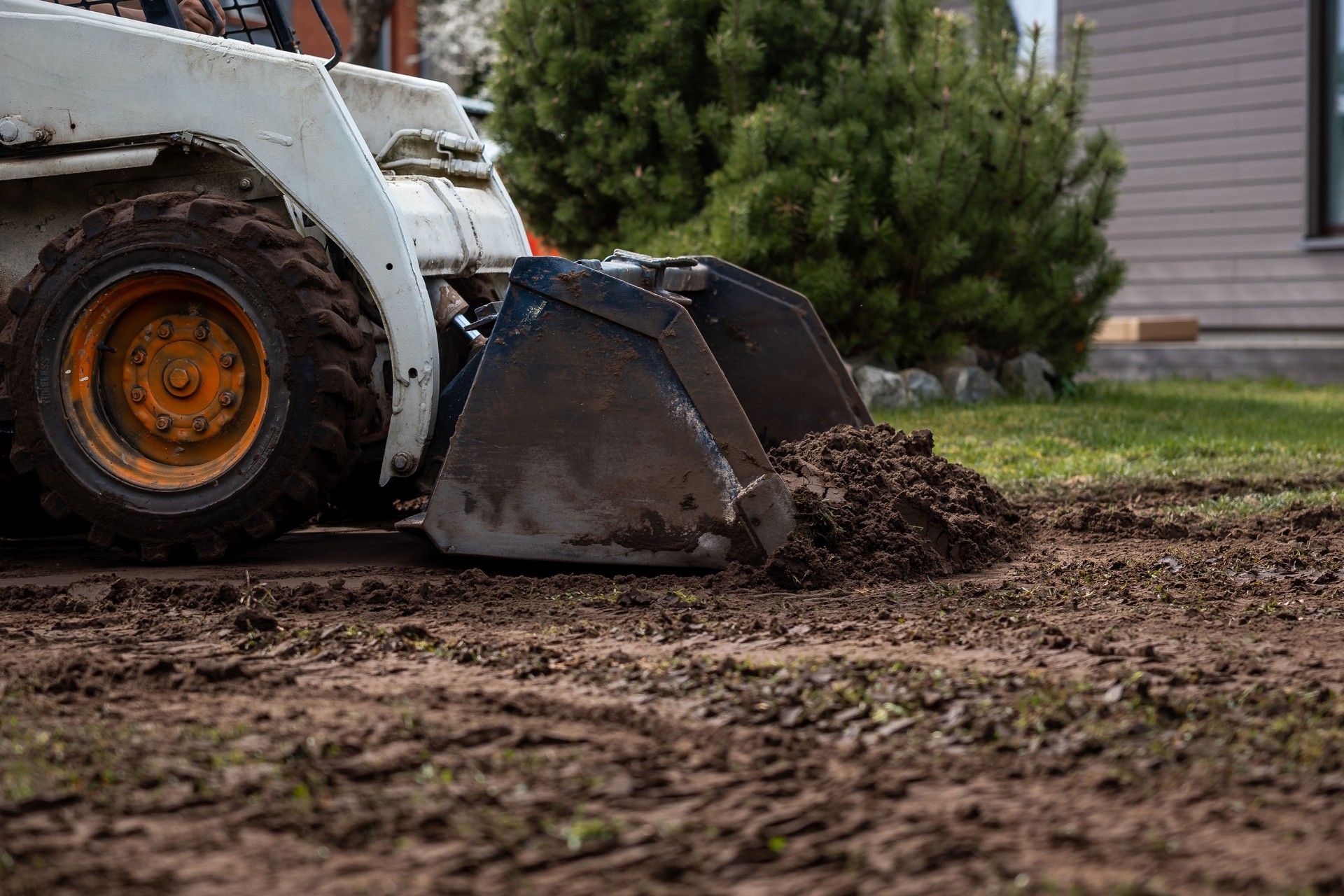 Skid Steer Loader Moving Soil Near Green Bush and Building